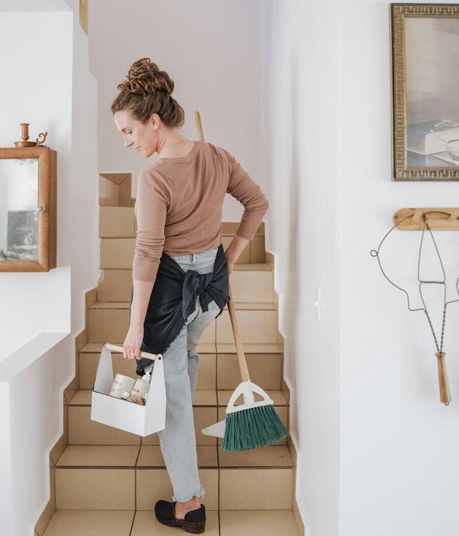 A cleaner carrying a caddy of supplies and a broom up a staircase